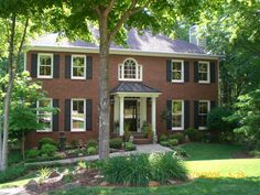 a large brick house with black shutters and white trim on the front door, surrounded by lush green trees