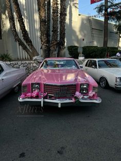 an old pink car is parked in front of some palm trees and other classic cars