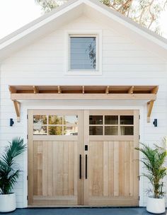 two wooden garage doors in front of a white house with potted plants on the side