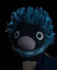 a black stuffed animal with blue hair and big eyes in front of a dark background