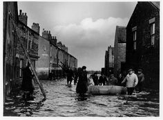 black and white photograph of people walking through flooded street with boat in water near buildings