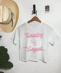 "Introducing our \"Country Legend\" Cowboy Crop Top - the perfect blend of rugged style and classic charm that's perfect for cowboys and country music enthusiasts alike. Whether you're two-stepping at your favorite honky-tonk or just want to showcase your love for the cowboy way of life, this crop top is the ideal choice. 🤠 Key Features: 🌟 Premium Quality: Crafted from soft and breathable fabric, this crop top ensures comfort and durability, making it ideal for everyday wear or special occasions. 🤠 Vintage Vibes: The timeless design features a bold \"Country Legend\" inscription in a rustic font, giving it a classic western feel. 🤠 Easy to Style: Pair it with your favorite jeans, boots, and a cowboy hat for the complete cowboy look, or layer it under a flannel shirt for a versatile out Country Style White Top With Graphic Print, Country Style White Cotton T-shirt, Country Style Cotton Tops With Letter Print, Country Style Short Sleeve Tops With Letter Print, Country Style Cotton Tops With Relaxed Fit, Crop Top Western, Country Music Outfit, Mode Country, Rodeo Outfit