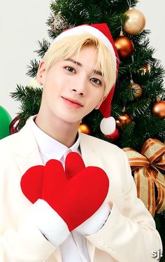 a man wearing a santa hat holding a red heart in front of a christmas tree