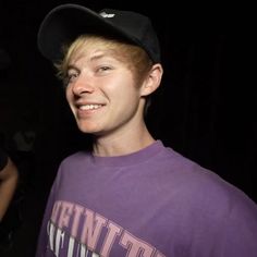 a young man in a purple shirt and black hat smiles at the camera while wearing a baseball cap