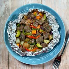 a blue plate topped with meat and veggies covered in tin foil next to a fork