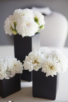 three black square vases with white flowers in them on a tableclothed surface