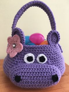 a purple crocheted purse with eyes and a flower on the handle sits on a wooden table