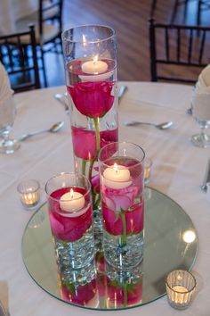 three tall vases filled with pink roses on top of a round table covered in candles