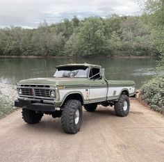 a green truck parked on the side of a dirt road next to a body of water