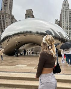 a woman standing in front of a large shiny object with people walking around it and buildings behind her