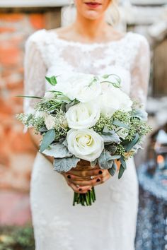 a bride holding a bouquet of white roses and greenery in her hands, standing next to a brick wall