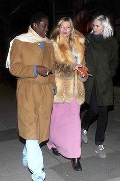 two women and a man are walking down the street at night, one is wearing a fur coat