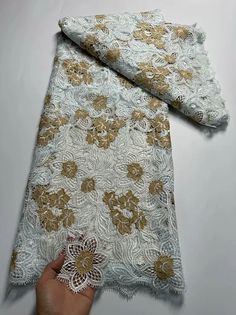 a hand is holding up a piece of white and gold lace with golden flowers on it