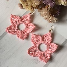 two pink crocheted flower shaped earrings sitting on top of a wooden table next to flowers