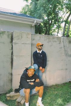 two men sitting next to each other in front of a wall and grass covered yard