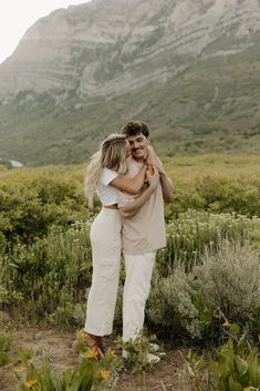 two people hugging each other in the middle of a field with mountains in the background