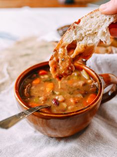 a hand holding a piece of bread over a bowl of soup with beans and vegetables