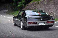 a black car driving down a road next to a lush green hillside covered in trees