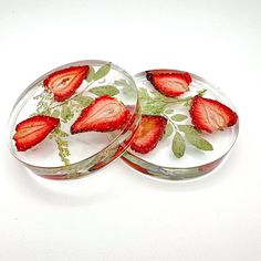 two glass plates with strawberries on them sitting on a white surface, one has green leaves and the other has red berries