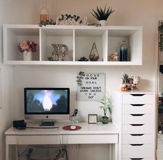 a white desk with a computer monitor and keyboard on it, surrounded by shelves filled with potted plants