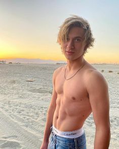 a shirtless young man standing on the beach at sunset with his hands in his pockets