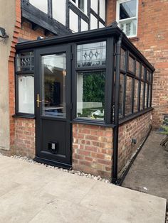 a small black building with glass windows on the outside and brick wall behind it, in front of a red brick house