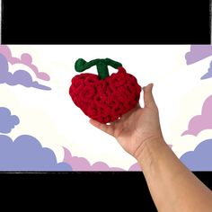 a hand is holding a red knitted strawberry ornament in front of a cloud background