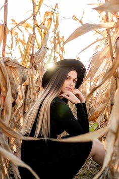 a woman with long hair sitting in a corn field wearing a black dress and hat