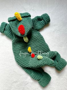 a green knitted toy laying on top of a white blanket with red, yellow and green pom poms