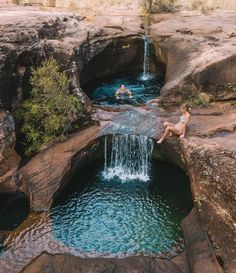 two people are swimming in a pool at the edge of a cliff near a waterfall