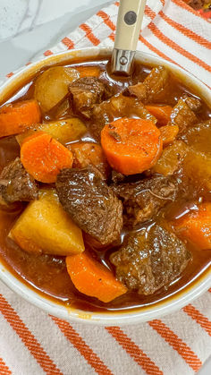 a bowl of beef stew with carrots and potatoes on a striped table cloth next to a spoon