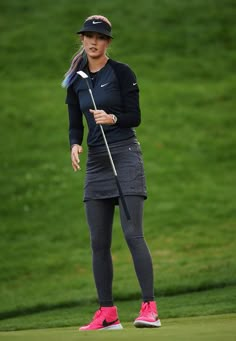 a woman holding a golf club on top of a green covered field with grass behind her