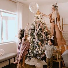 two children and an adult decorating a christmas tree in a living room with balloons