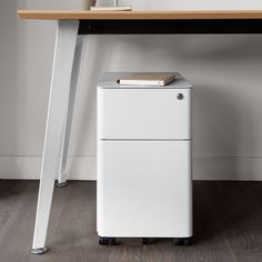 a desk with a book on top and a white trash can underneath it, in front of a wooden table