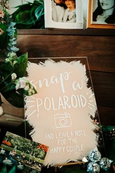 a polaroid sign sitting on top of a table next to flowers and pictures with the words snap a polaroid and leave a note for the happy couple