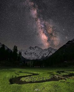 the night sky is filled with stars above mountains and green grass, as well as some water