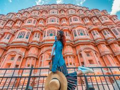 a woman in a blue dress and straw hat walking past a tall pink building with balconies on it