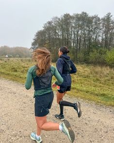 two people running on a dirt road near some trees and grass, with one person in the background