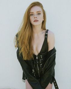 a woman with long red hair wearing a black leather outfit and posing in front of a white wall