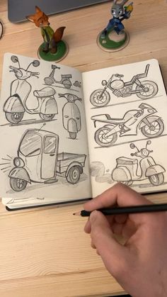 a hand holding a pencil over an open book with cartoon drawings on it and various toy motorcycles