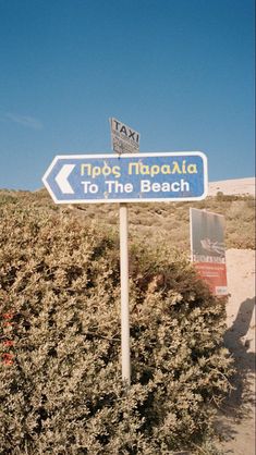 a sign pointing to the beach on top of a hill with bushes in front of it