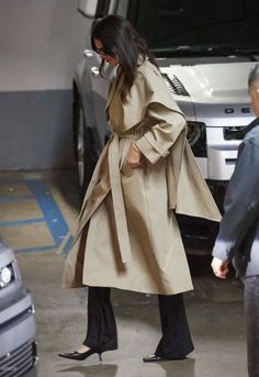 a woman in a trench coat and black pants is walking towards a silver car with her hand on her hip