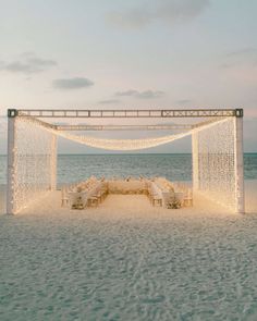 an outdoor wedding set up on the beach with white linens and lights strung over it