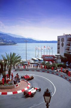 two race cars driving around a curve on a road next to the ocean with people watching