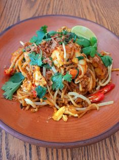 a brown plate topped with noodles and vegetables on top of a wooden table next to a lime wedge