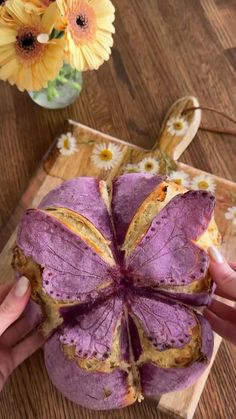 a person holding up a purple flower shaped pastry on a cutting board with flowers in the background