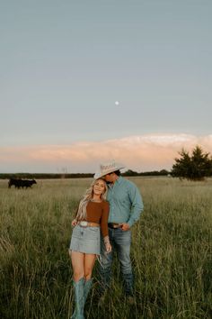 a man and woman are standing in the tall grass with cows behind them at sunset