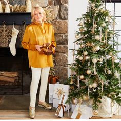 a woman standing next to a christmas tree with presents in front of her and stockings on the fireplace