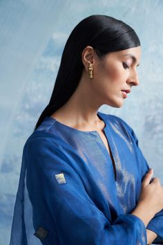 a woman with her arms crossed wearing a blue dress and gold earrings, standing in front of a wall