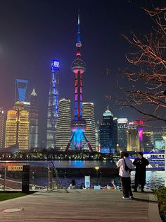 two people are standing on a dock in front of the city skyline at night time